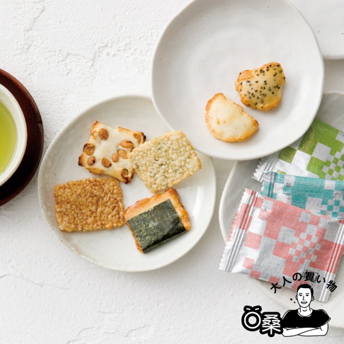 這張俯視角度的圖片展示了多種不同口味的日式米果。左下方盤子盛放著四種米果，包含表面鑲嵌豆子的方形米果、酥脆質地的長方形米果、灑滿細碎顆粒的米果，以及包覆一片海苔的米果。右上方盤子則有兩片圓弧狀米果，其中一片點綴著黑色芝麻。右側疊放著三包包裝精緻的鋁箔袋裝米果，整體呈現淡雅的日式茶點風格。