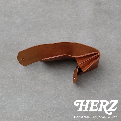 一個採用淺棕色皮革製作的HERZ職人手作迷你短夾，呈現展開狀態，露出內部多層次的零錢收納隔間與金屬扣環設計，背景為灰色水泥紋理，右下角印有品牌標誌與產地資訊。