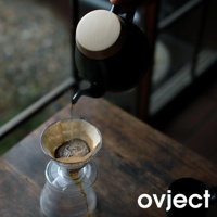 黑色琺瑯手沖壺，搭配木質壺蓋，正從壺嘴傾倒熱水至咖啡濾杯中，濾杯內有咖啡粉與水滴，下方為玻璃分享壺。