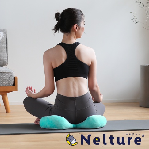 一位女性身穿運動服，背對鏡頭，盤腿坐在一個藍綠色的瑜珈坐墊上，坐墊下方是一張瑜珈墊，圖片右下角有日文和英文的品牌標誌。