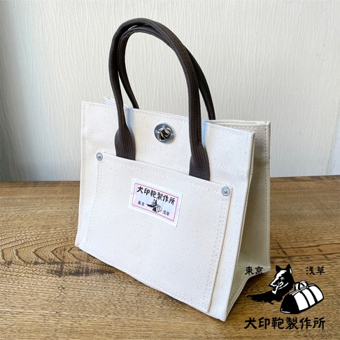 犬印純棉帆布迷你托特包，米白色帆布材質，配有深棕色提把。包包正面有一個大口袋，上面貼有帶有日文圖案的標籤，並有金屬鈕扣固定。