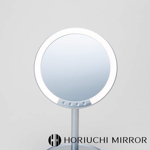 銀色金屬材質的桌上型化妝鏡，圓形鏡面外圈有LED燈，鏡面下方有三個觸控按鈕，鏡子架設在一個金屬支架上，背景為淺灰色。