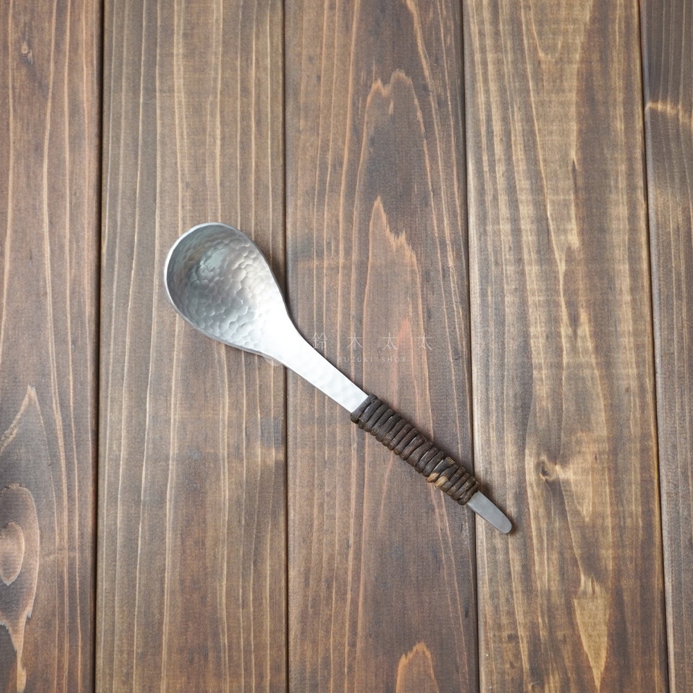 銀色鋁製湯勺，勺面帶有錘目紋，勺柄由深棕色編織材質包裹。