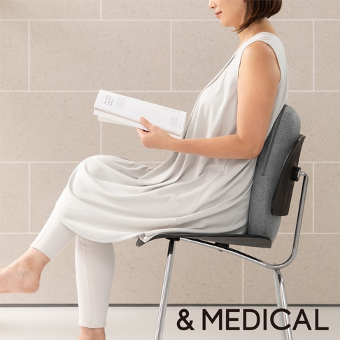 一位女士坐在椅子上，手持一本白色雜誌，背部倚靠著一個灰色布製的肩頸背按摩指壓枕，枕頭側面有黑色塑膠支架。