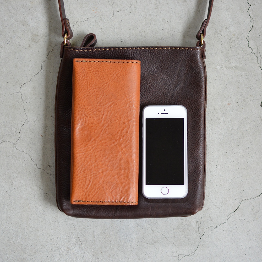 深棕色皮革製成的肩背包，包面上擺放著一個淺棕色長夾和一支白色手機，展示包的容量與尺寸。
