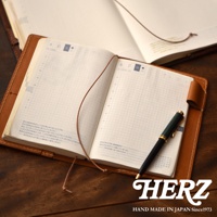 一本職人手作的棕色皮革手帳書套，內頁為方格筆記本，書套旁有一支綠色與金色的鋼筆，背景為木紋桌面。