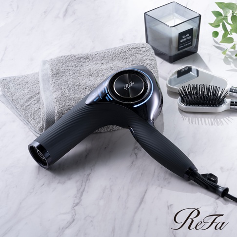 ReFa BEAUTECH DRYER PRO 智能吹風機，黑色機身，設有圓形控制面板，周圍帶有藍色指示燈。機身帶有紋理，配有灰色毛巾、鏡子和黑色髮刷。