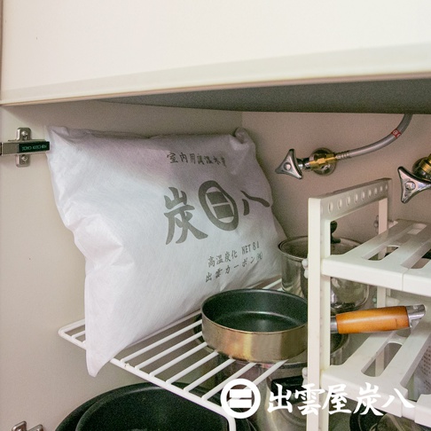 一個白色包裝袋，印有灰色日文文字「炭八」，標示「室內用調濕木炭 NET 8L」，袋子置於白色層架上，層架旁有鍋具和金屬水管。