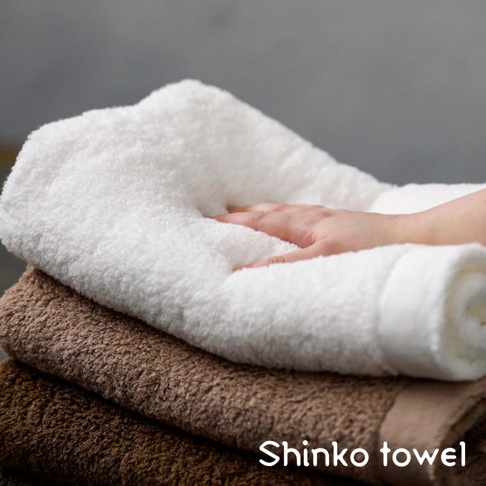 MAISON BLANC 輕奢柔膚匹馬棉浴巾，疊放展示。頂層為白色浴巾，中間有一隻手輕柔觸碰，下方為兩條棕色系浴巾。