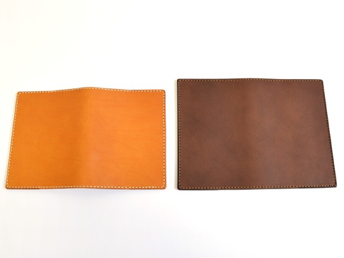 兩個職人手作皮革厚革書套，一個為原色皮革，另一為深棕色皮革，皆以白色縫線邊緣裝飾，展現皮革的自然紋理與手工質感。
