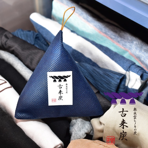 深藍色立體三角形調濕炭包，表面有網格紋理，頂部有金色掛繩，附有標籤，標籤上有「古來炭」字樣及繩結圖案。產品置於衣物堆中。
