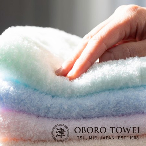 一疊三條毛巾，由上至下顏色分別為淺綠、淺藍、淡紫。毛巾材質蓬鬆柔軟，一隻手正輕柔地觸摸最上方的毛巾。毛巾質感細膩，呈現出高質量。