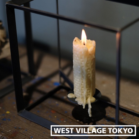黑鐵製長方展示架內，點燃一支滴蠟的黃色蜂蠟柱狀蠟燭，蠟燭表面有蜂巢紋理，火焰明亮。