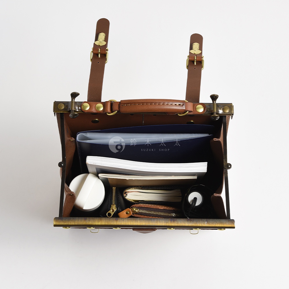 職人手作皮革復古口金後背包，棕色皮革與古銅色金屬配件，內部裝有文件夾、筆記本、化妝包和白色圓形物體。