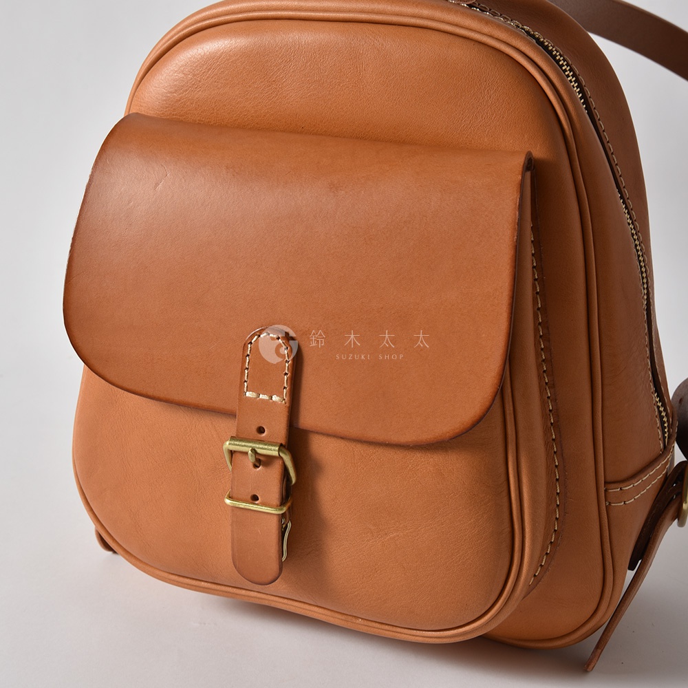 職人手作的淺棕色皮革迷你後背包，具有立體前袋，以銅扣和皮革帶固定，縫線清晰。