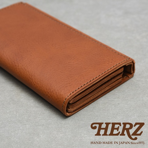 一張從高角度拍攝的日本HERZ職人手作皮革長夾特寫，棕色皮革紋理細緻，展現出厚實且富有質感的裁切邊緣與細膩的縫線細節。圖片右下角印有HERZ品牌標誌及HAND MADE IN JAPAN Since1973字樣，背景為簡約質感的灰色桌面。
