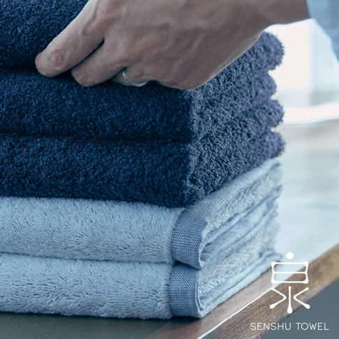 一疊深藍色和淺藍色的柔織有機棉浴巾，其中一疊浴巾由一隻手拿起，浴巾邊緣有細緻的織紋。