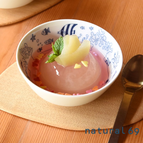 白色陶瓷小碗盛裝著粉色果凍，頂部放著水煮梨片與薄荷葉，碗內裝飾著彩色糖珠。碗身印有藍色海洋生物圖案，碗置於木質托盤上，旁有一支金色湯匙。