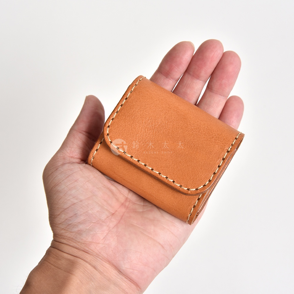 一隻手掌托著一個小巧的棕色皮革零錢包，零錢包表面帶有天然皮革紋理，採用淺色粗線手工縫製而成，外型為雙折設計，邊緣處理細緻。