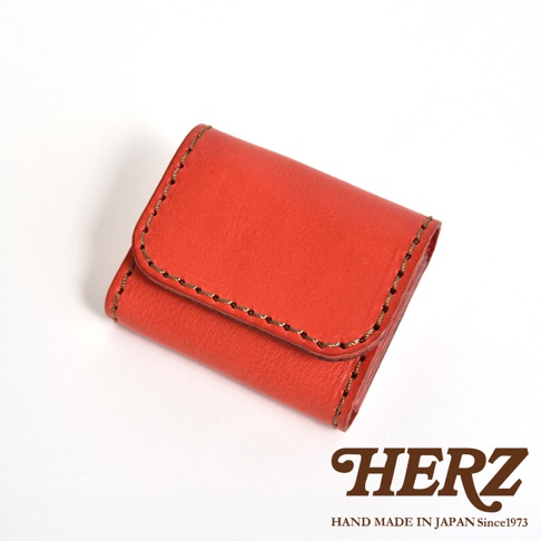 一個精緻的日本HERZ職人手作紅色皮革雙折零錢包，表面呈現自然的Stare皮革紋理，邊緣以深色粗線手工縫製，整體呈現簡約耐用的設計風格，右下方印有HERZ品牌標誌。