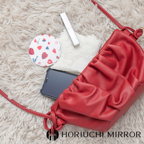 NAPURE plune. 隨身口袋小圓鏡，圓形鏡面飾有紅色和藍色愛心圖案，搭配白色蕾絲、紅色皮革包、手機和銀色唇膏，散落在絨毛地毯上。