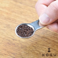 手指握持著一個由18-8不鏽鋼製成的銀色雙頭計量咖啡豆勺，其中一個勺頭裝滿了細緻的咖啡粉末，勺子放置於木紋背景上。