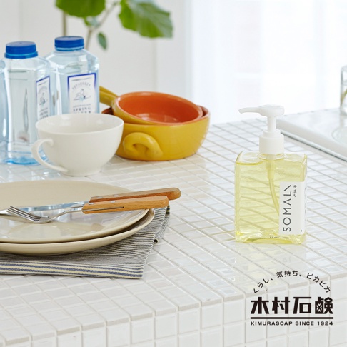透明方形壓瓶裝有淡黃色液體，瓶身標有「SOMALI」字樣。背景為白色磁磚檯面，擺放有餐盤、餐具、白色杯子、黃色與橘色陶碗，以及兩瓶藍色瓶蓋的礦泉水。