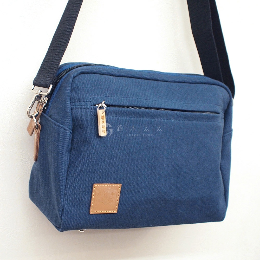 一個藍色斜背包，帆布材質，有一個拉鍊前袋，上面有一個木製吊牌，上面印有日文文字，包包底部有一個方形皮革補丁，上面有壓花圖案。