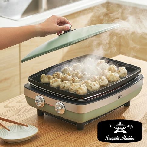 綠色帶金屬邊框的電烤盤，盤中擺放著許多煎餃，正冒著熱氣。一隻手正提起淺綠色的鍋蓋，鍋蓋邊緣有黑色把手。烤盤下方有兩個銀色旋鈕，烤盤本體為綠色和米白色。