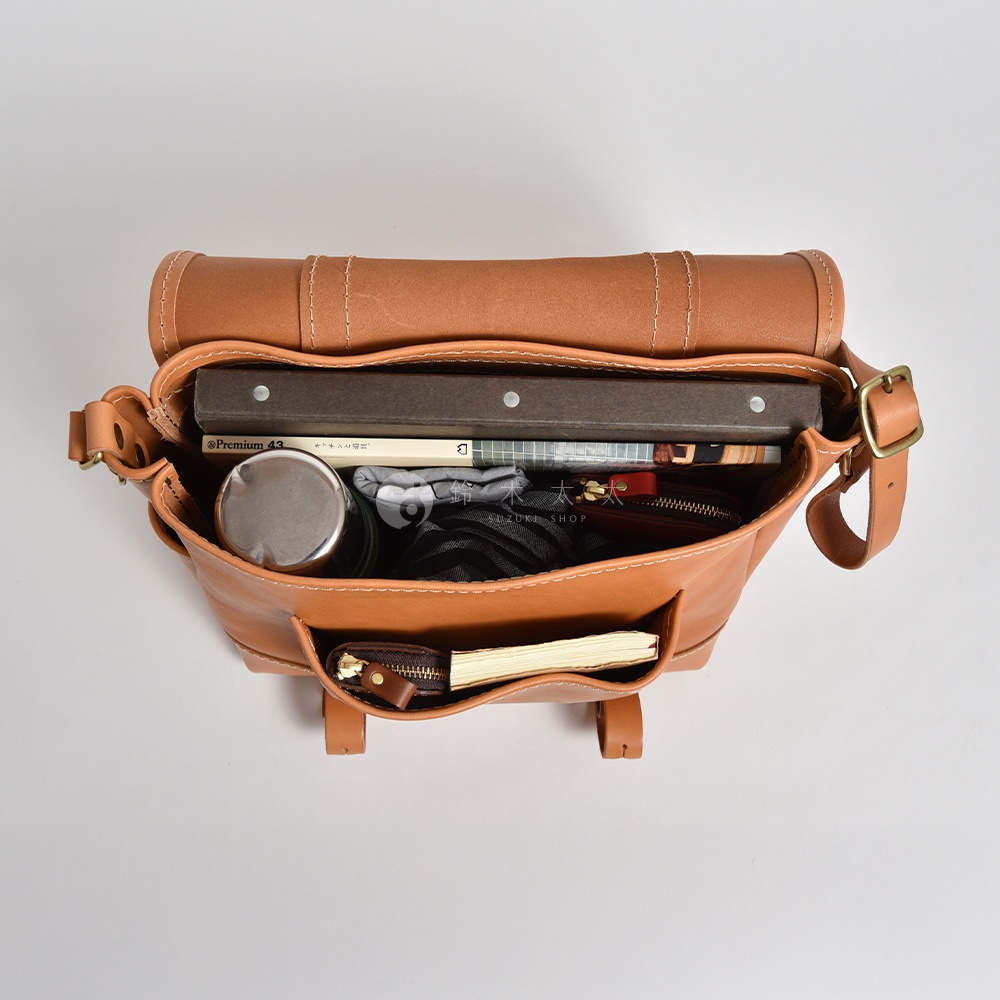 職人手作皮革復古郵差包，棕褐色牛皮材質，頂部視角展示包內裝有書籍、保溫杯、錢包及筆記本，金屬扣環和車縫線清晰可見。