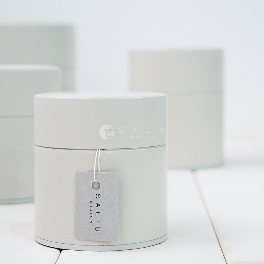 淺灰色圓柱形錫製茶葉罐，罐身簡約無飾，蓋子上方印有品牌標誌，側邊掛有灰色吊牌，吊牌上印有「SALIU DESIGN」字樣。