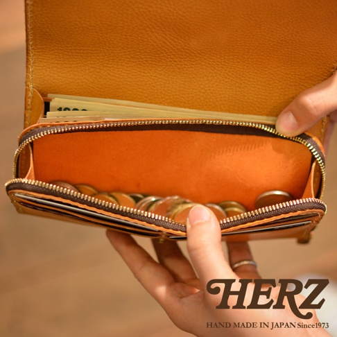 焦糖色皮革長夾，內部為亮橘色，拉鍊開合，裝有紙鈔與硬幣，展現寬口設計與大容量。