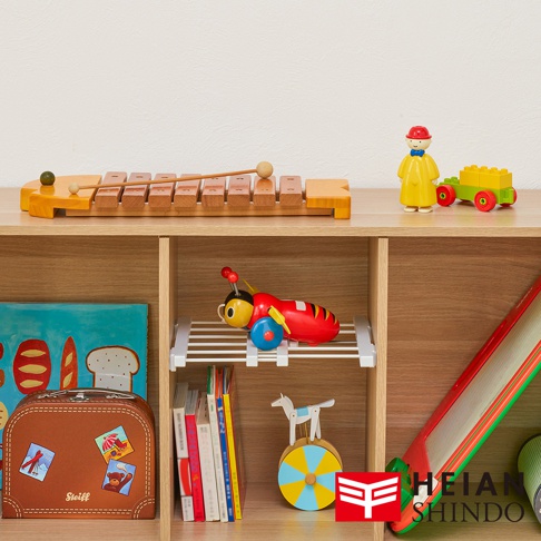 層架上擺放著多種玩具，包含一個木製的木琴、一個紅色小蜜蜂造型的玩具、一組黃色小人偶和綠色小火車，以及疊放的書籍和一個棕色行李箱造型的置物盒，右側還有一個大型紅色滑梯和品牌標誌。
