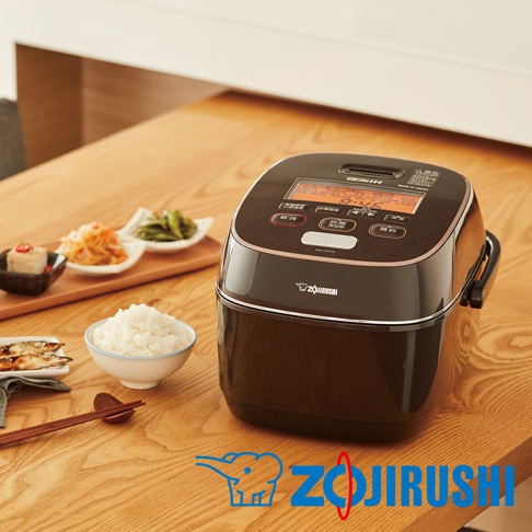 一台深棕色的鐵器豪熱羽釜壓力IH電子鍋，配有數位顯示螢幕和多個按鈕。鍋身光滑，搭配銀色飾邊。旁邊擺放著一碗白米飯、幾碟日式小菜和烤魚，以及一雙筷子。