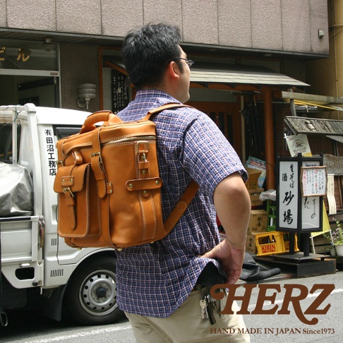 職人手作皮革大型三用旅行後背包，採用棕色皮革，設計有頂部提把、拉鍊開口、多個前袋及側邊收納空間，搭配金屬扣環和車縫線細節，適合旅行及日常使用。