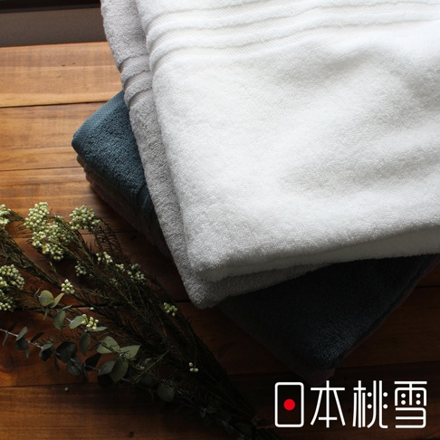 三條今治飯店浴巾堆疊，分別為白色、淺灰色和深藍色。浴巾材質為柔軟的棉，表面有細緻的條紋織法。左側擺放著一束乾燥的綠色植物作為裝飾，背景為木紋桌面。