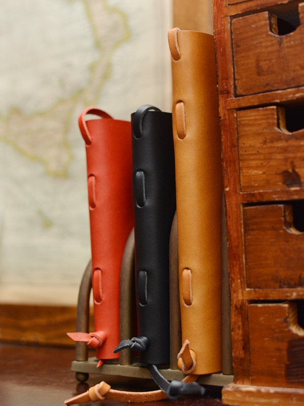 三款職人手作皮革附繩書套，分別為鮮紅色、黑色及原色皮革，皆有頂部和側邊的皮革環扣設計，底部有皮革繩結，整體呈現簡約風格。