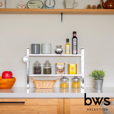 bws SELECTION 不鏽鋼可伸縮雙層收納架，架上擺放著各種罐裝食品、調味料瓶、玻璃罐以及一個編織籃，左側有一個木碗裝著紅色蘋果，背景為淺色牆面和木質櫃檯