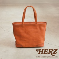 職人手作皮革Organ肩背拉鍊托特包，採用橘棕色皮革，簡約設計，手提把為雙層結構，包身底部有加固皮革，右下角有HERZ品牌字樣和HAND MADE IN JAPAN Since1973文字。