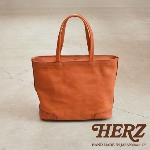 職人手作皮革Organ肩背拉鍊托特包，採用橘棕色皮革，簡約設計，手提把為雙層結構，包身底部有加固皮革，右下角有HERZ品牌字樣和HAND MADE IN JAPAN Since1973文字。