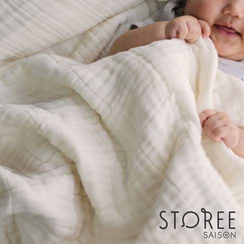 象牙白色九層紗單人毯，質地輕柔，表面有菱格紋縫線。嬰兒的手和臉部露出毯子一角。