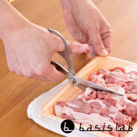 一雙銀色不鏽鋼萬用料理剪刀，左手正握住剪刀，右手扶著裝有生肉的橘色塑膠盒，剪刀的刀刃正在切割肉塊。