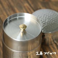 銀色不鏽鋼錘目紋茶葉罐，配有金色圓形提手，罐身有明顯的錘擊紋理，另一同款茶葉罐蓋子在背景中模糊顯示。