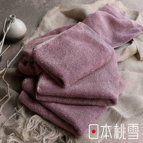 一疊粉紫色精梳棉飯店毛巾，觸感柔軟，質感細緻，部分毛巾疊放，部分攤開，背景為淺色粗織布料與灰色石紋表面，左側有白色裝飾枝幹與陶瓷罐。