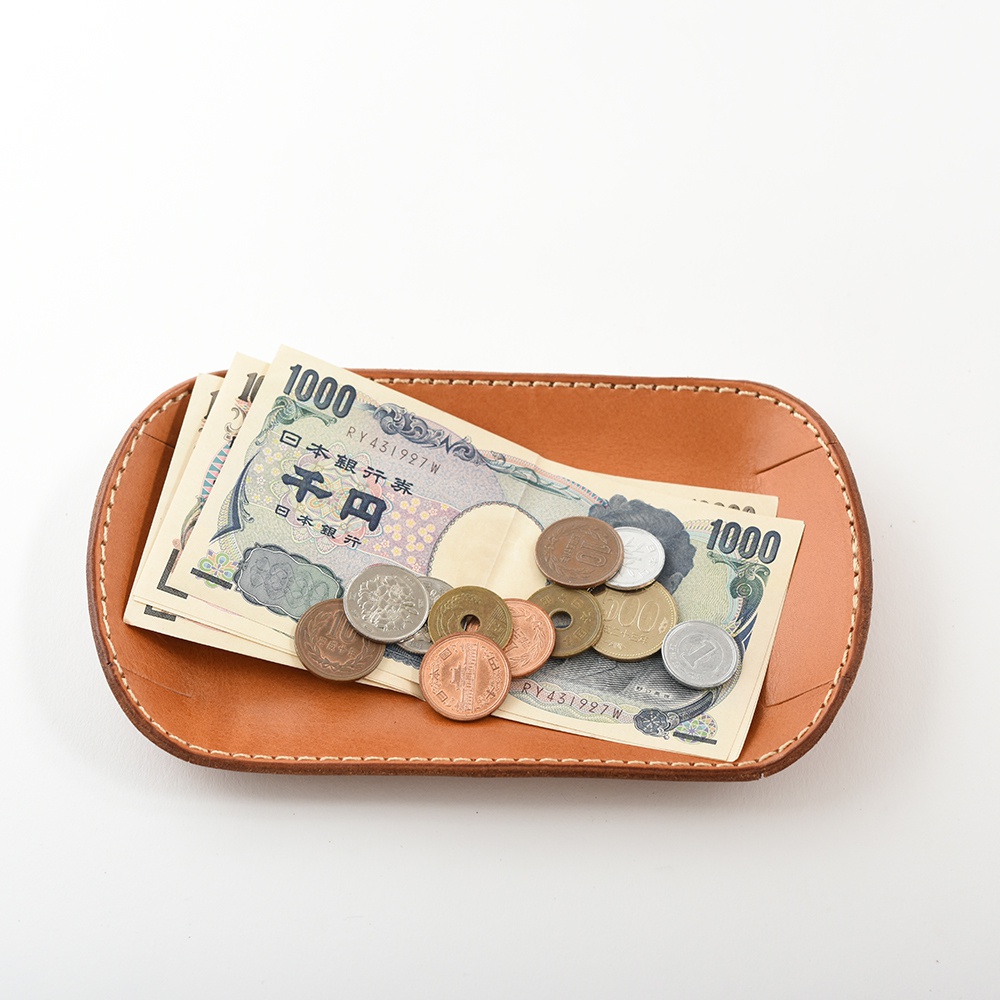 職人手作皮革小物收納盤，淺棕色皮革，盤中放置多張日圓紙鈔及硬幣，盤邊緣有縫線裝飾。