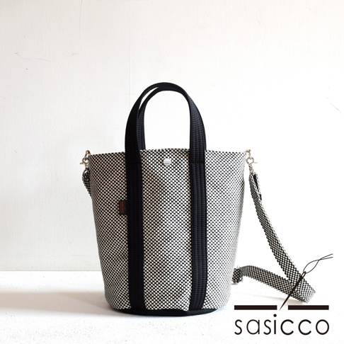 三河木綿刺子織兩用水桶包，棋盤格紋黑白色，搭配黑色織帶提把與可拆卸斜背帶，包身結構挺立。