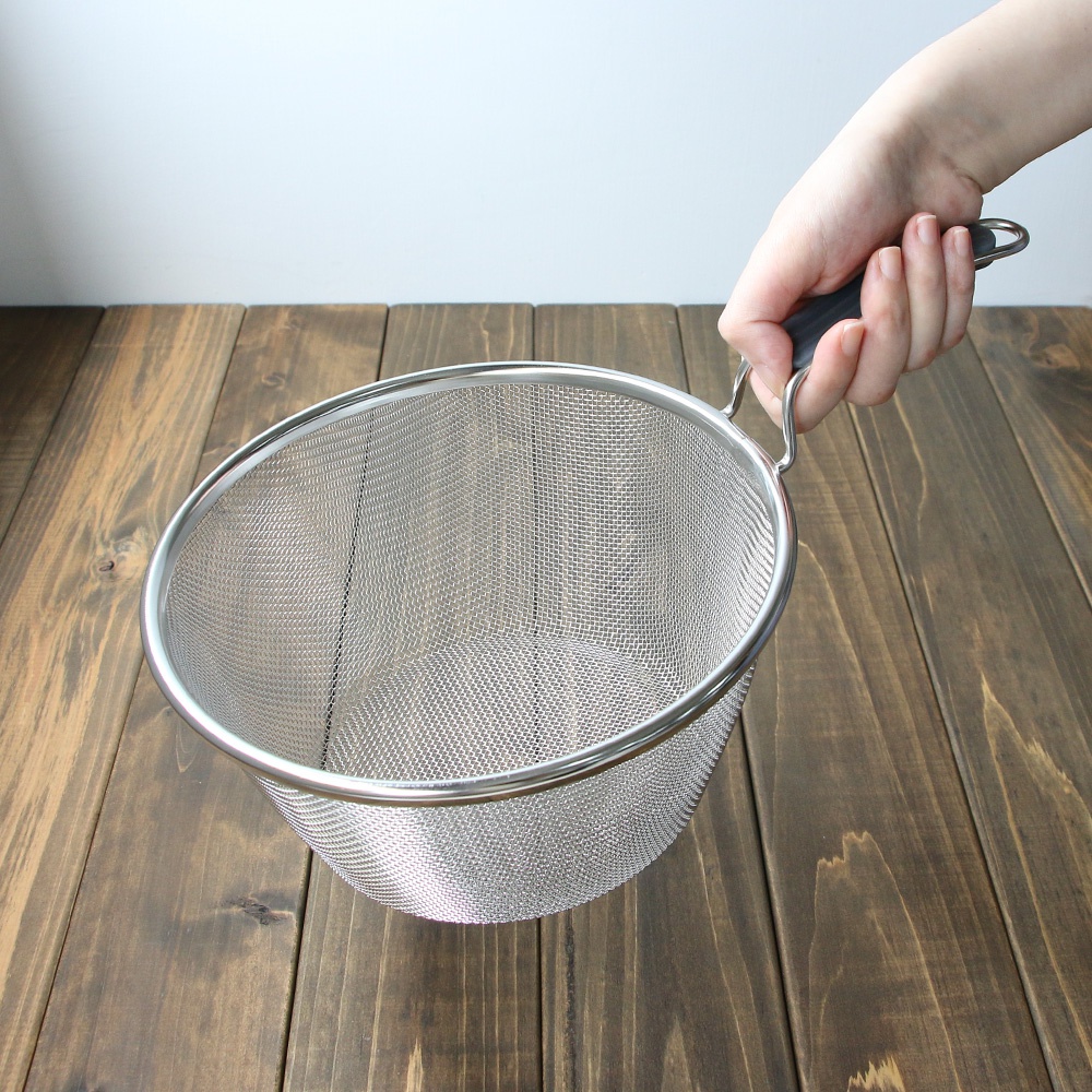 手持一個 20 公分的不鏽鋼濾網，濾網為細網狀，邊緣和提把為銀色不鏽鋼，握柄處為黑色矽膠材質。