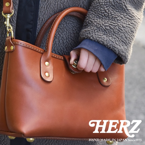 職人手作皮革迷你托特包，採用棕色皮革製成，配有雙提把、金屬旋轉鎖扣和可拆卸肩帶。包包底部有四個金屬腳釘，增添質感。