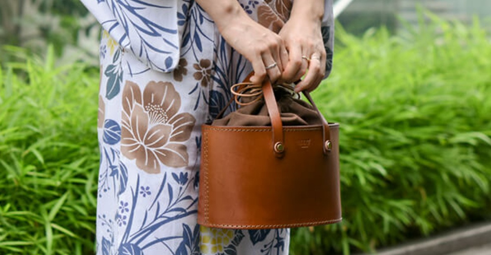 棕色皮革束口手提包，包身呈長方形，配有同色系皮革提把，包內為深棕色布料束口設計。