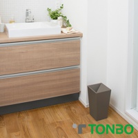 一個灰褐色的方形半開垃圾桶，放置在木質地板上，旁邊是木紋櫥櫃及白色洗手台。